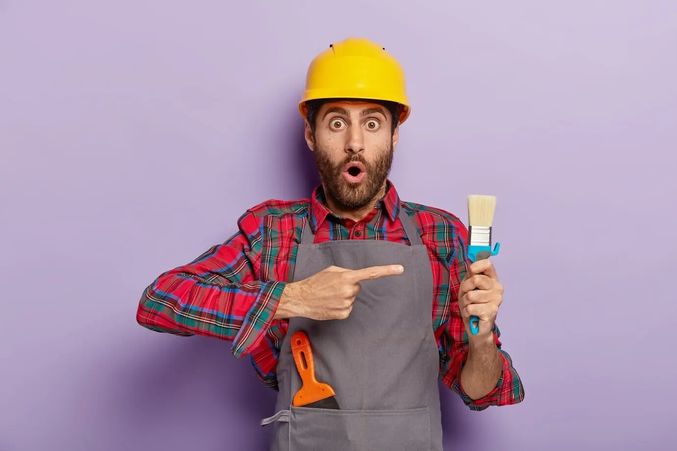 Trabajador manual estupefacto señala una herramienta de reparación, ocupado en trabajos de reconstrucción, lleva casco y uniforme especial. Trabajador de la construcción sorprendido muestra una brocha de pintar mientras está en el trabajo. Renovación.